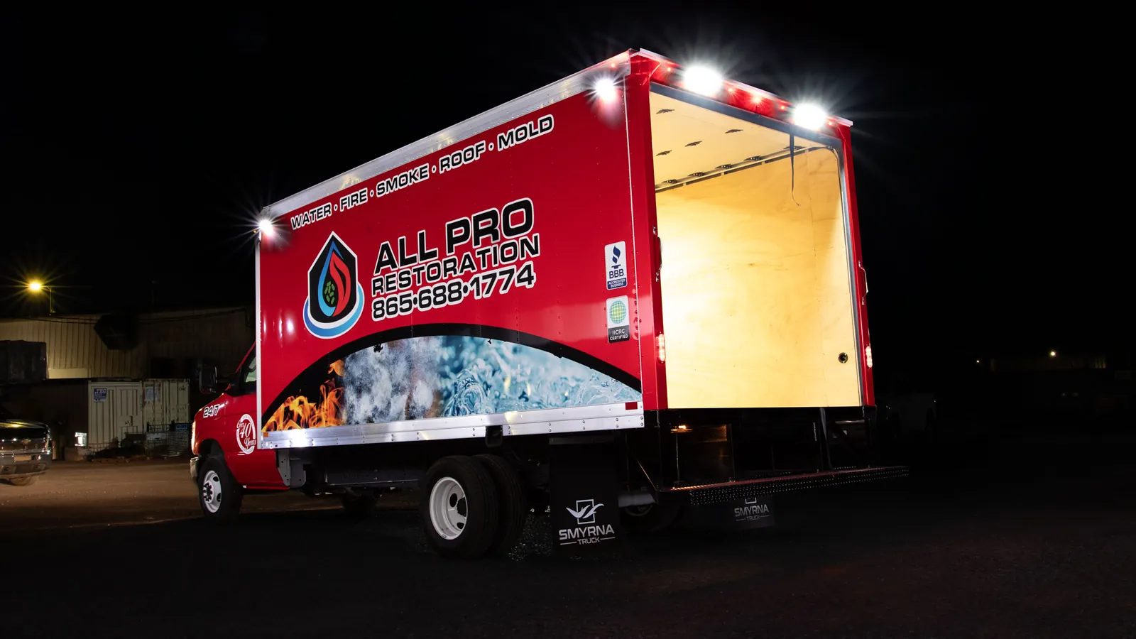 The same box truck with the rear door open, showing the lit plywood-lined cargo area and roof work lights